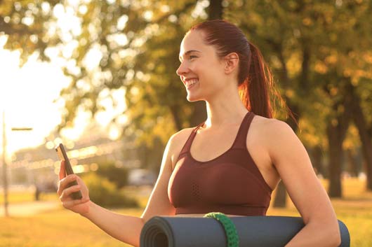 Cute smiling woman wearing tracksuit, using smartphone and holding yoga mat
